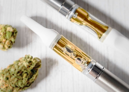 Filled vape cartridges rest on a white table next to some cannabis buds