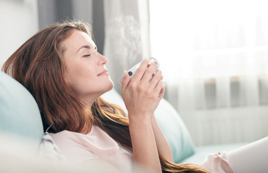 A young woman is smelling a cup of coffee with her eyes closed. She is sitting on the couch